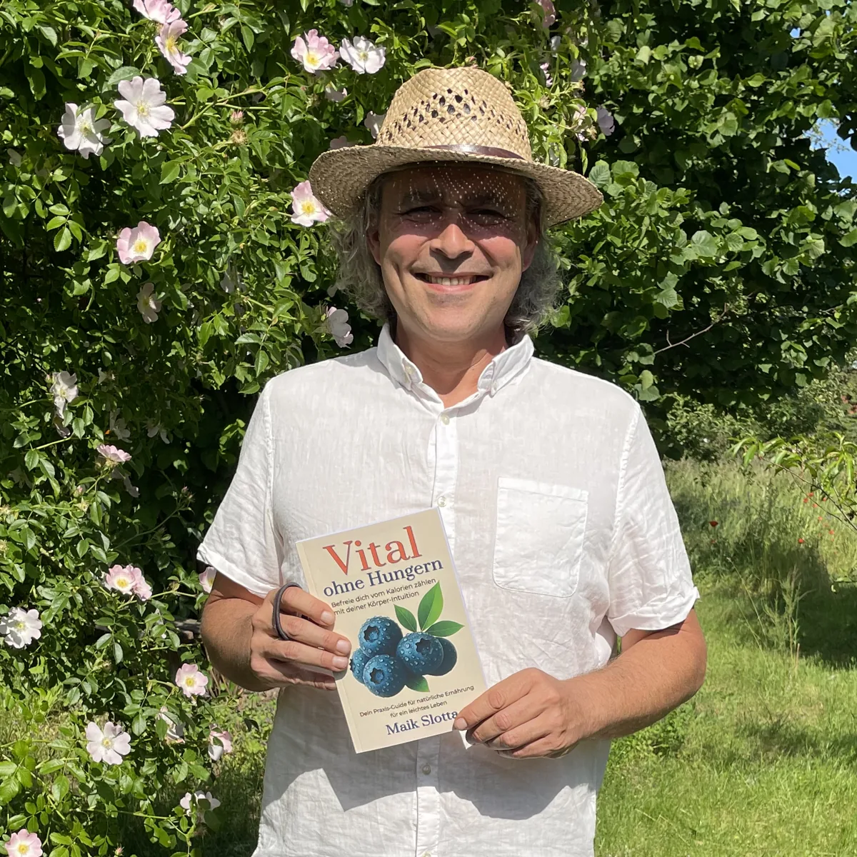 Maik mit Strohhut hält sein Buch Vital ohne Hungern vor einem blühenden Rosenbusch