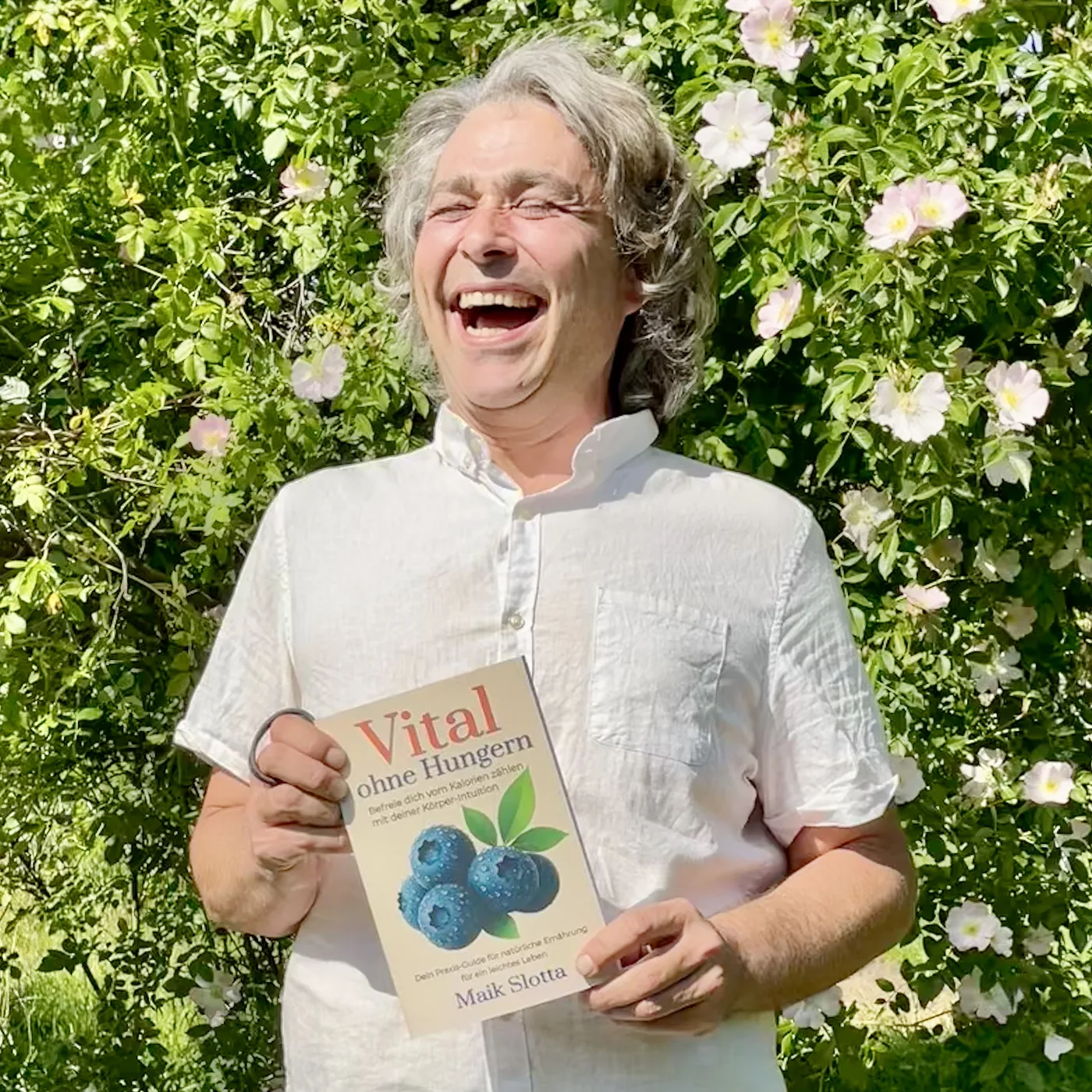 Maik lacht herzlich und hält sein Buch Vital ohne Hungern vor blühenden Wildrosen